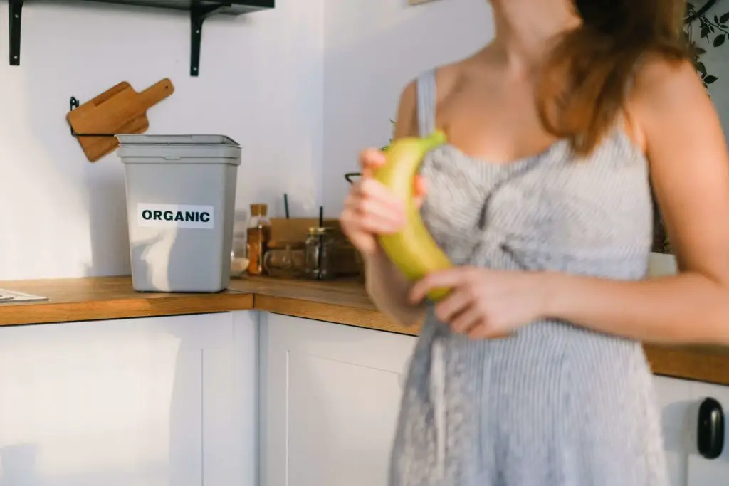 A woman is standing holding a banana in front of her compost bin, after she eats the banana, the peel will go into the bin for organic waste.