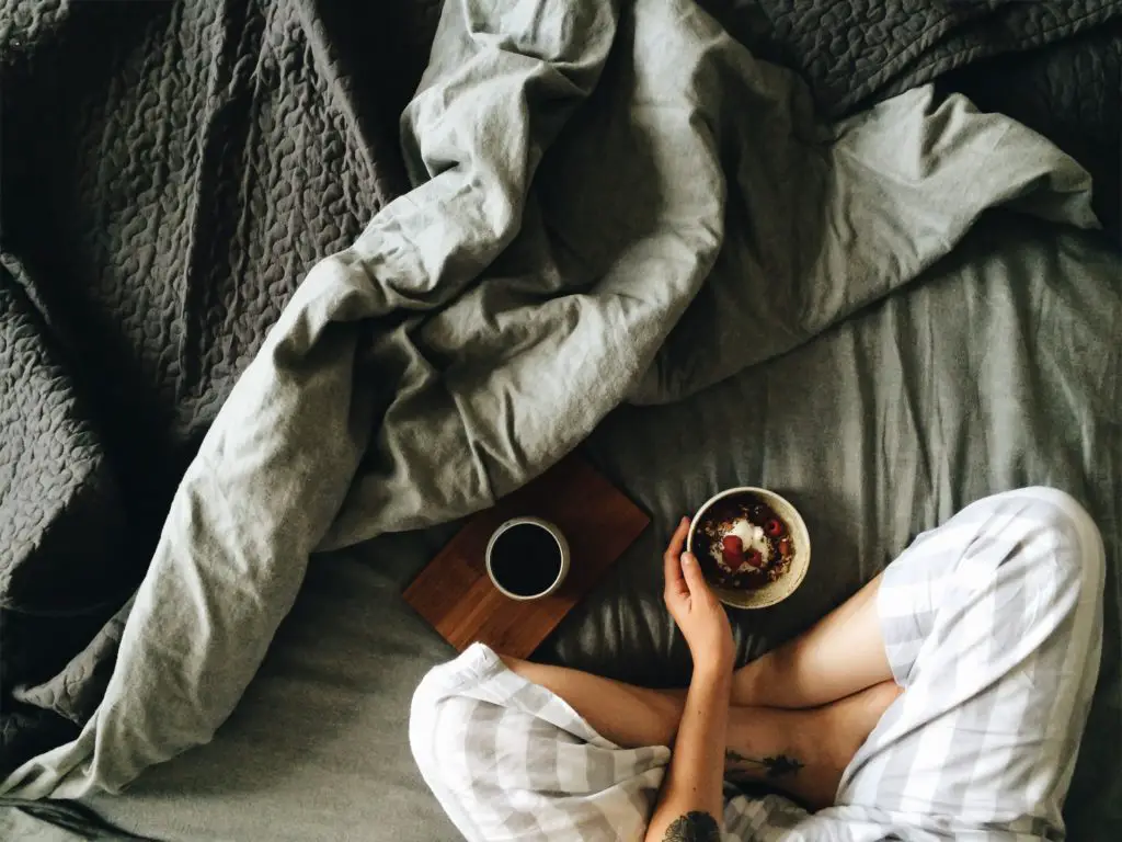 This photo shows a woman embracing the joy of missing out (JOMO) as she relaxes in bed with a cup of coffee.