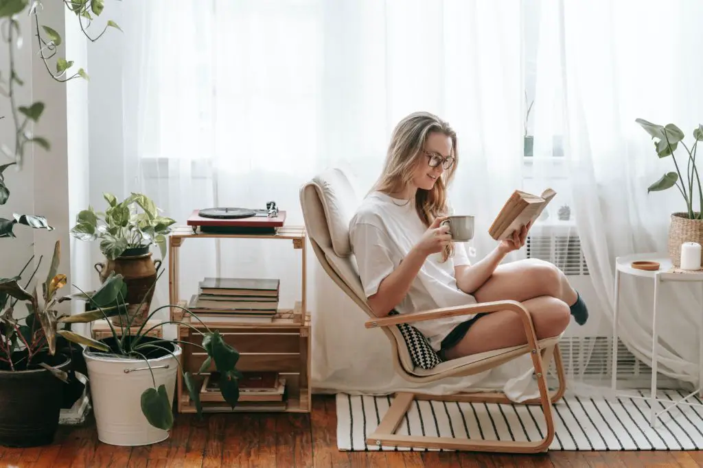 This photo of a woman reading while sipping coffee, sitting on a big comfy chair, surrounded by plants, shows what plants can add to the space.