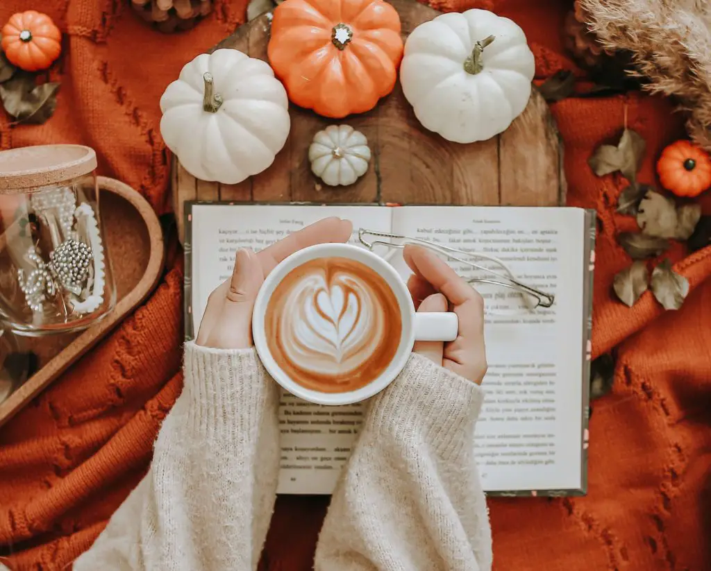 A warm and cozy latte held by hands wearing a knitted sweater, featuring fallen leaves and small pumkins. 