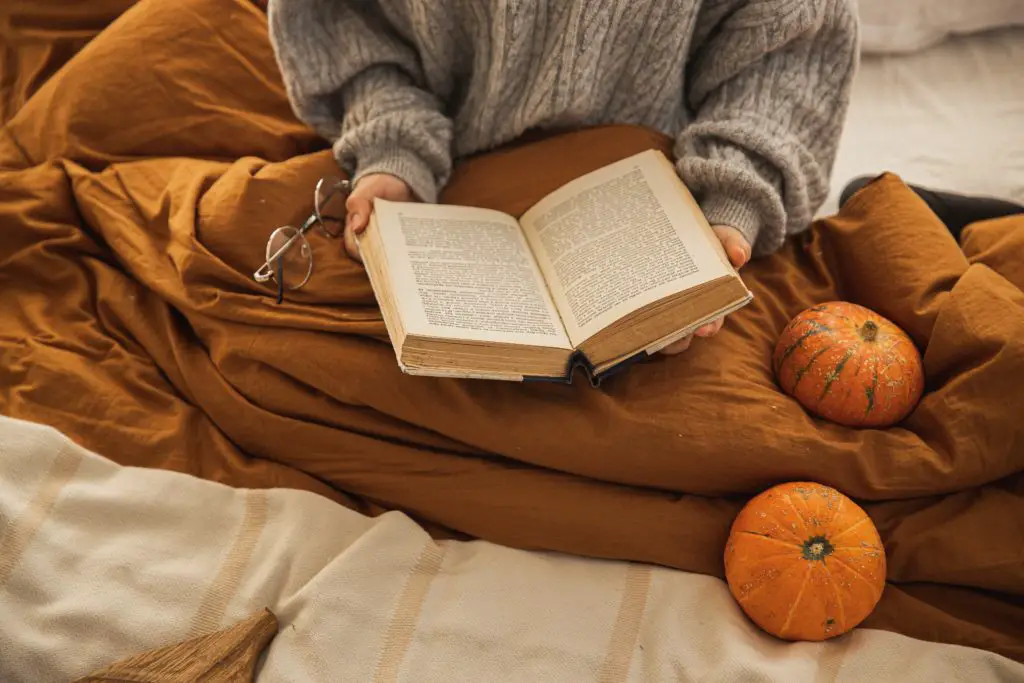 A woman sitting amongst tiny pumpkins and a cozy blanket reading fall poetry.